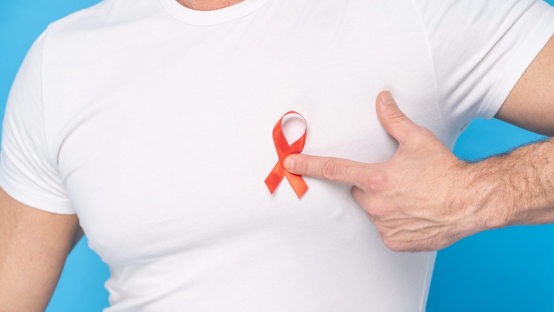 Man hold red ribbon bow AIDS awareness symbol on a heart wearing a white t-shirt isolated on a blue background. Modern medicine and healthcare. AIDS awareness concept.
